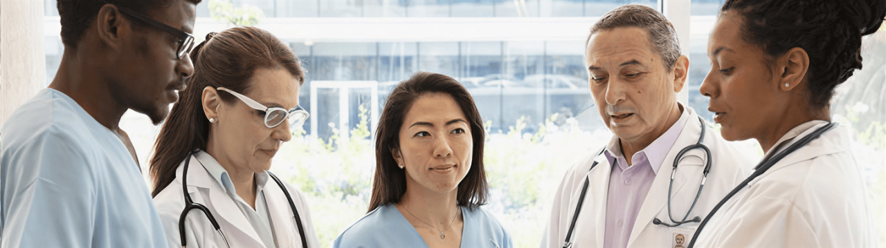 A group of five medical professionals in lab coats and scrubs stand together in a bright, modern healthcare setting, engaged in a focused discussion.