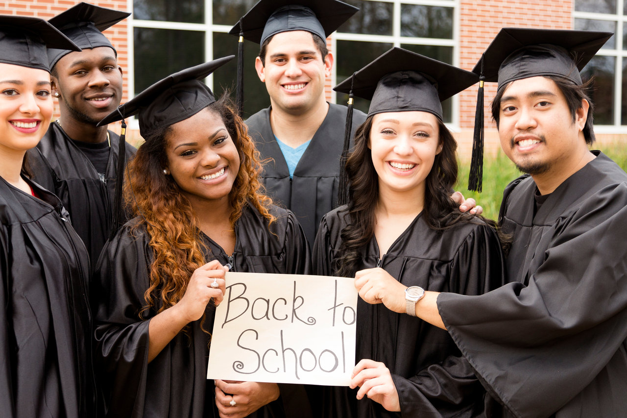 Six, multi-ethnic friends dressed in black cap and gowns excitedly hold "Back to School" sign after college graduation. University school building background.