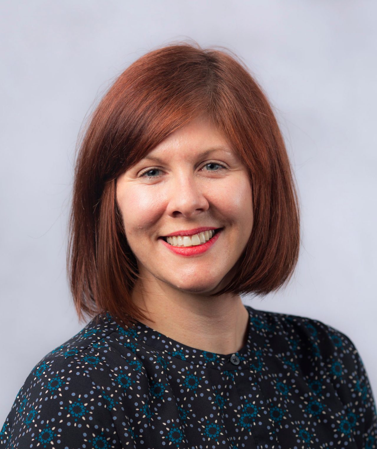 white woman with red hair, red lipstick, with navy shirt with turquoise circles and white dotted circles 