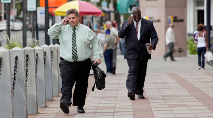 two overweight men talk on their cell phones while walking down a city street