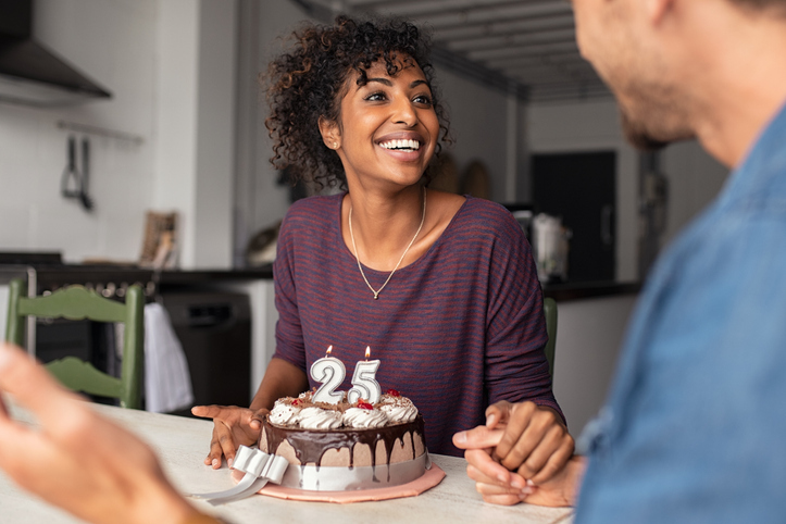 Multiethnic couple sitting at table and celebrating birthday at home in living room. Young african girl surprised on seeing 25th birthday cake with her boyfriend. Excited wife celebrating birthday with husband at home.