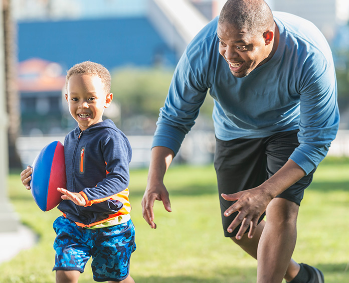 Black man in blue shirt and black shorts chasing black boy with red and blue football