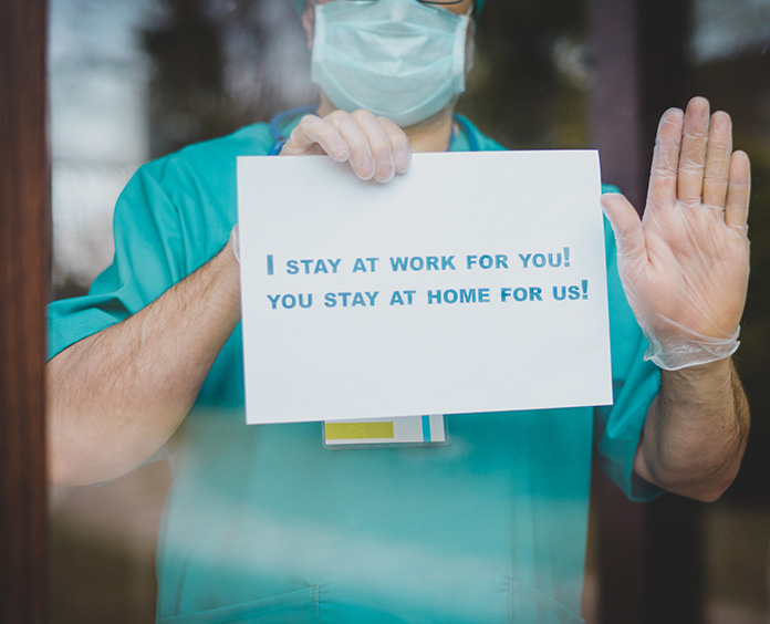 person in aqua shirt holding up one hand like a stop and a sign in other that says I stay at work for you! You stay at home for us! 