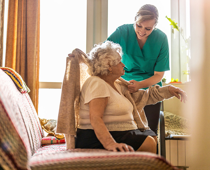 Gray haired black large woman seated getting helped with sweater by younger woman in green medical shirt