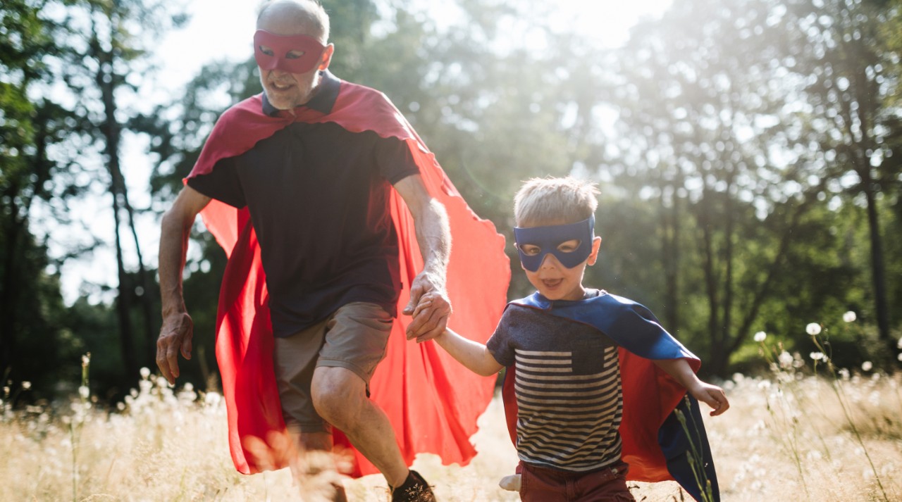 A senior man has fun dressing up in a hero mask and cape costume with his grandchild, a boy about 4 years old.  They have fun playing a running around in a sunny grassy field.