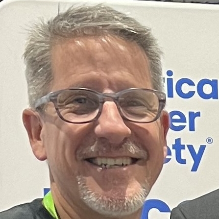 White man with brown hair, fluffy bangs, gray thin beard and moustache