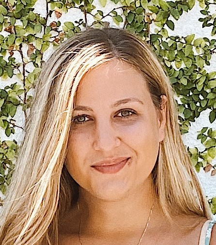 headshot of woman with long straight red hair parted on right in front of  green bushes