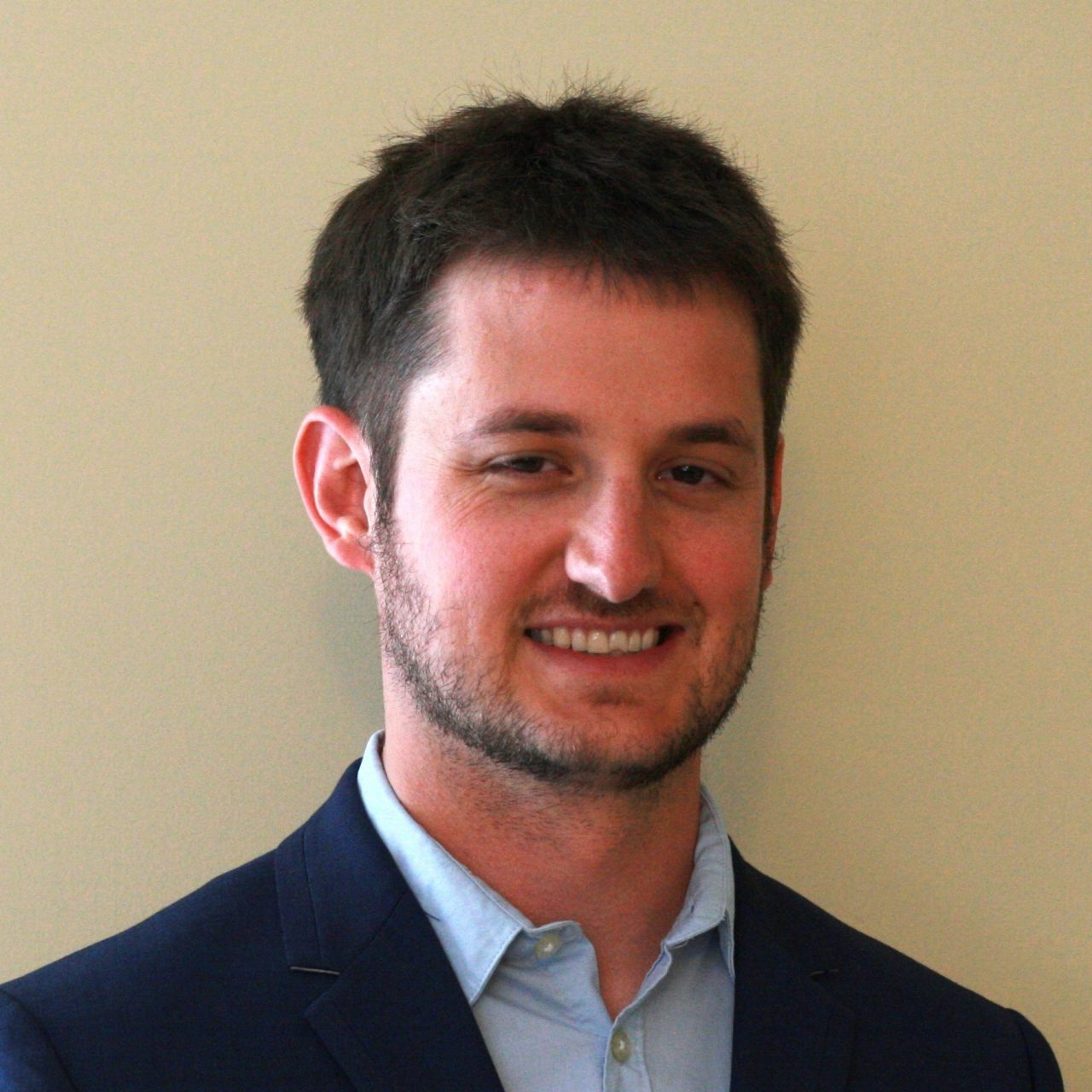 white man against beige wall wearing dark gray jacket, white shirt, and red and blue tie